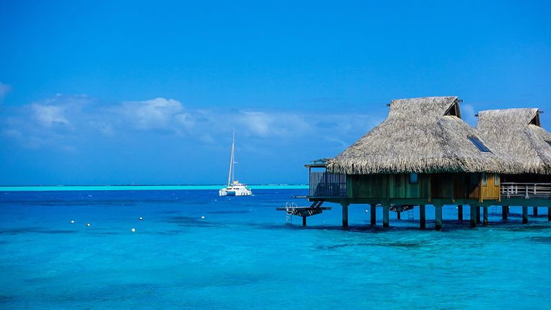 Overwater bungalows in blue water in Bora Bora with catamaran in background against blue sky